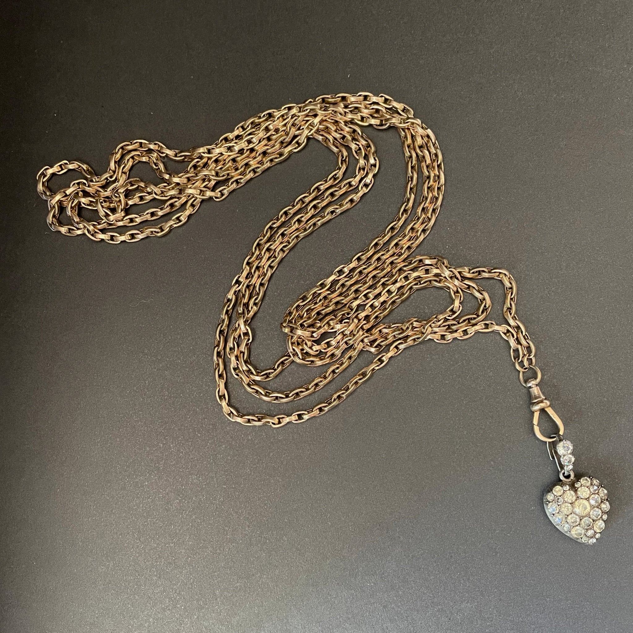 Gold long guard chain with swivel clasp holding a heart shaped pendant with colourless stones.