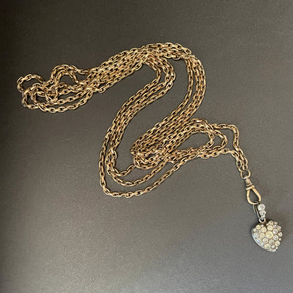 Gold long guard chain with swivel clasp holding a heart shaped pendant with colourless stones.