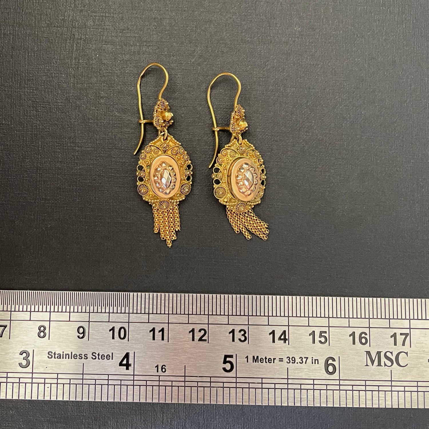 Gold drop earrings on a black background with a metal ruler.