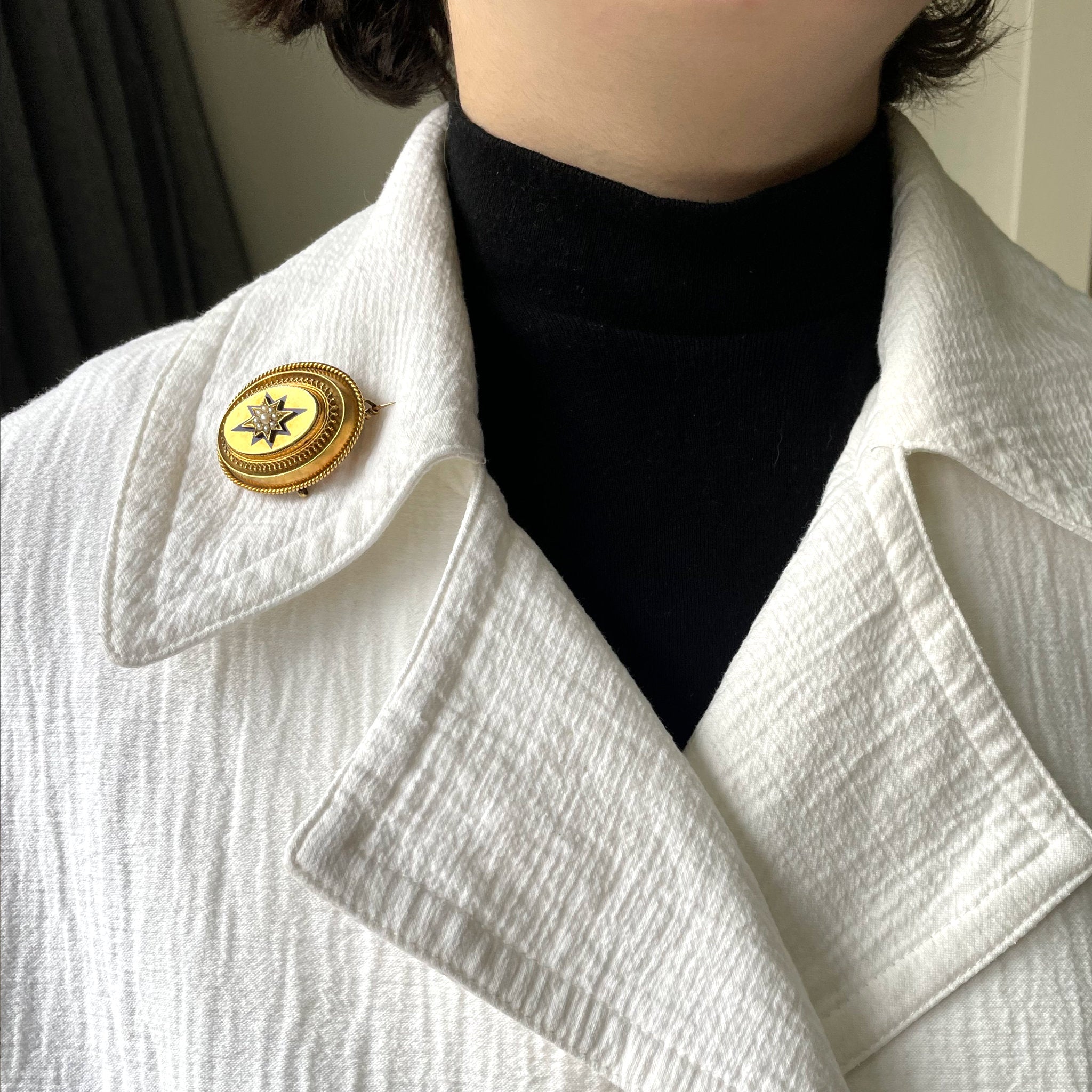Woman wears large oval gold locket with pearls and white jacket.